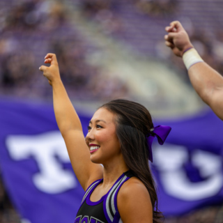 A smiling TCU Showgirl makes the two-fingered "Go Frogs" hand sign at a crowded football game.