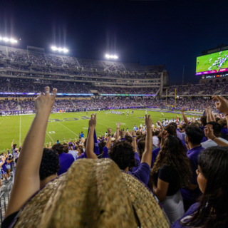 Dozens of cheering TCU fans raise their arm in a two-fingered "Go Frogs" hand sign at a crowded football game.