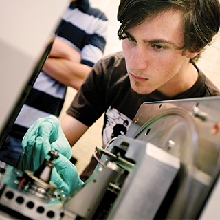 College of Science & Engineering A male student wearing green protective gloves works on a piece of electronics