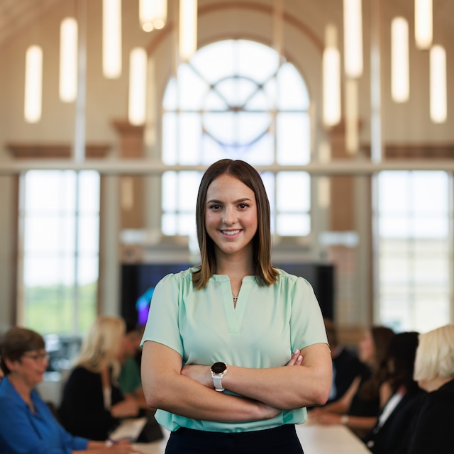 A student stands with her arms folded in front of a long table of people