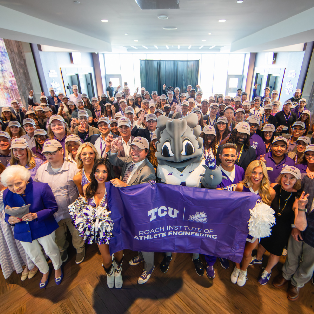 A group of individuals with TCU mascot SuperFrog holding a flag of the Roach Institute of Athlete Engineering